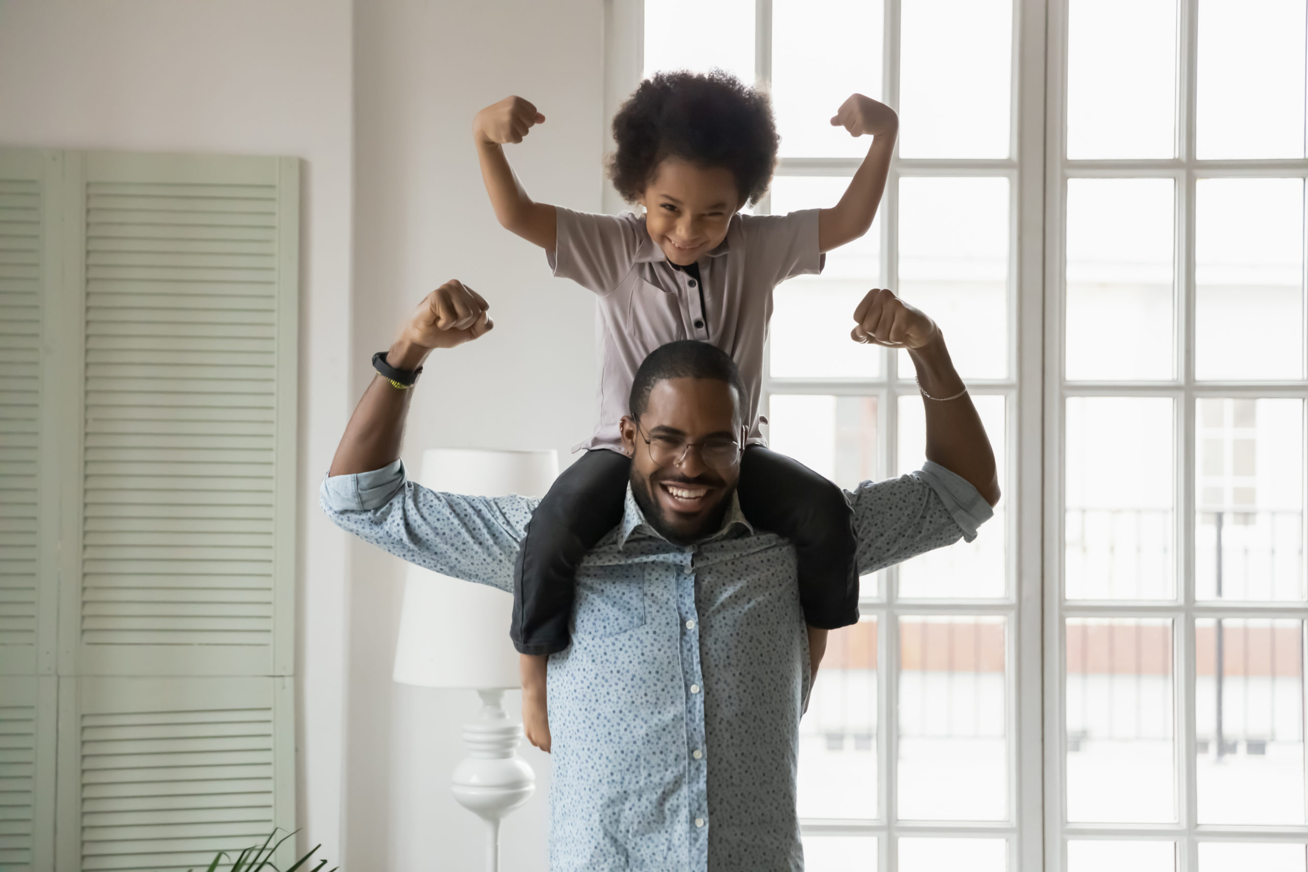 African ethnicity little son sitting on fathers shoulders showing biceps