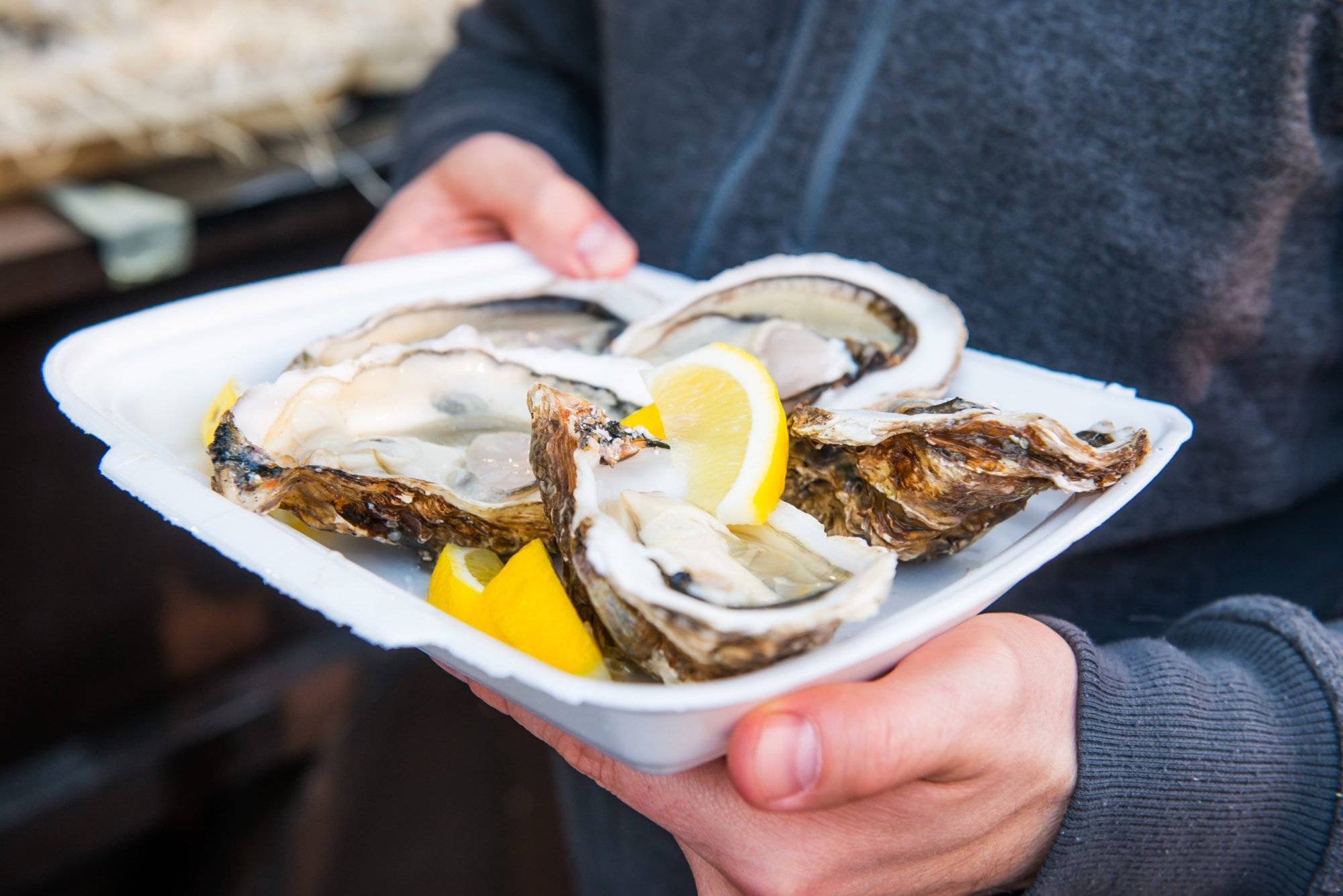 Close up male hand holding take away food tray with fresh opened oyster and lemon slices at street food market, festival, event. Selective focus. copy space.