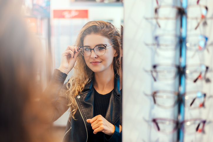 Smiling young woman trying on glasses on mirror in optician.